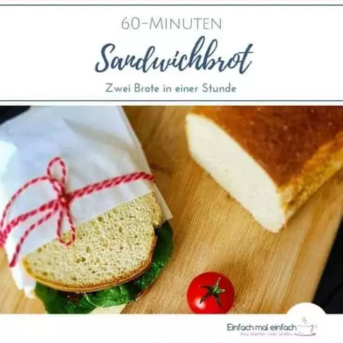 Weißes Kastenbrot auf Holzbret mit Sandwich in Butterbrottüte und rotem Garn zugebunden. Tomaten, Salat und dunkle Soße auf dem belegten Brot.